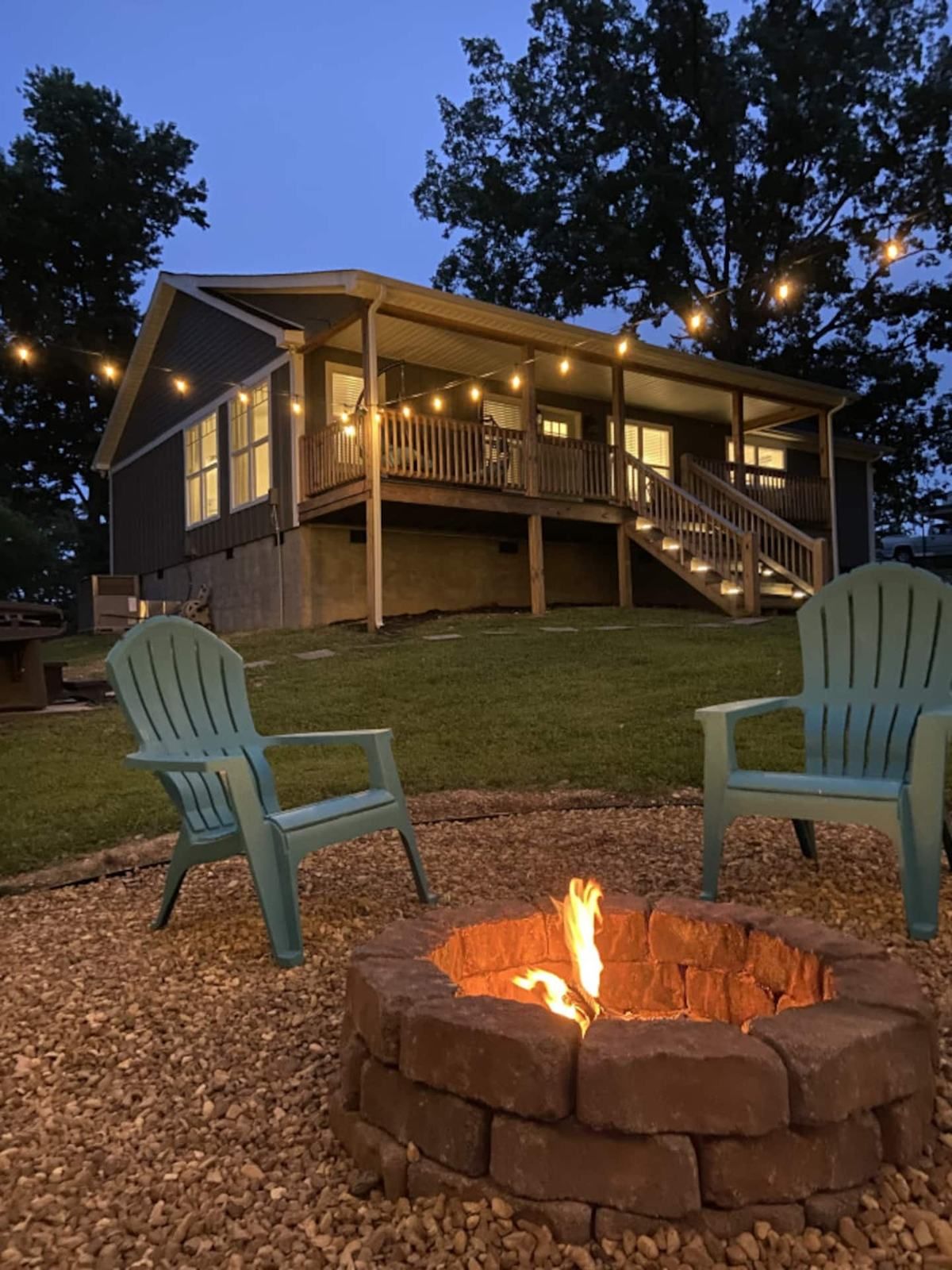 The house at dusk, string lights on the porch and a fire burning in the stone fire pit ringed by Adirondack chairs