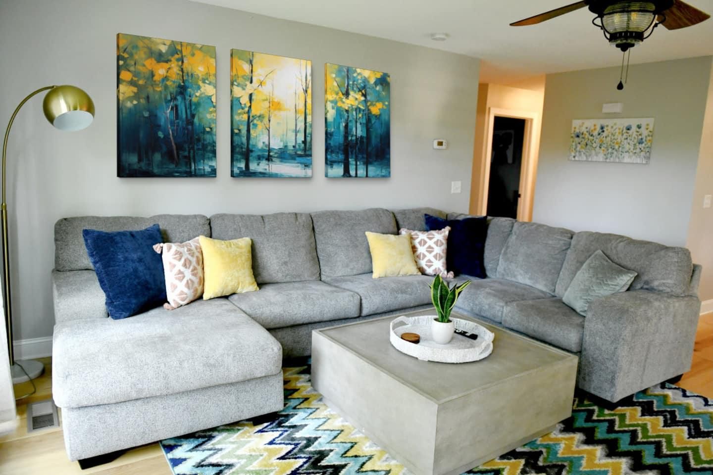 Living room with a large grey sectional, forest paintings and a patterned rug